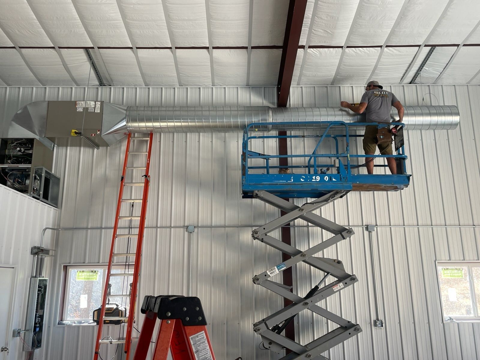 A Rockley Mechanical technician on a scissor lift installing spiral ductwork along a metal building wall